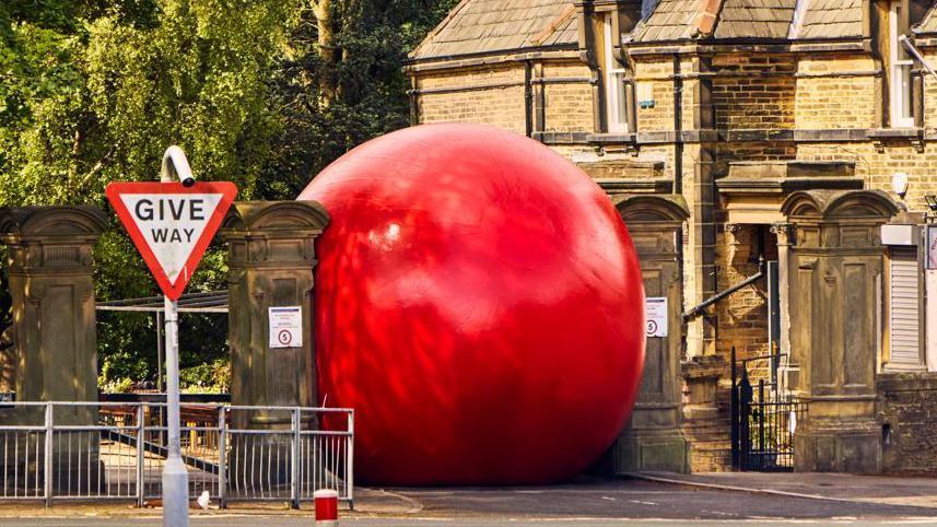 A large red ball is wedged between two stone pillars. Railings are infront of the ball to the left and ahead of those is a Give Way sign.