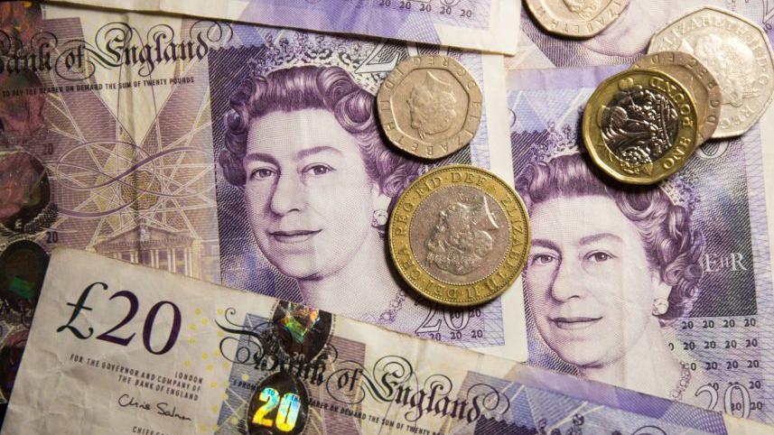 A pile of £20 notes with the face of Queen Elizabeth II. There are also coins scattered on top.