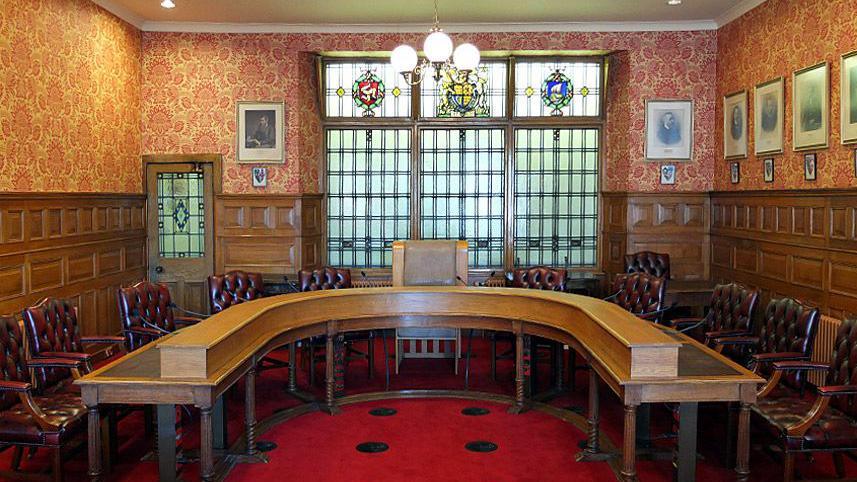 The Legislative Council chamber, which has wooden panelled walls and features a horseshoe shaped wooden table with chunky leather clad chairs. There are portraits of former governors on the wall on the right and large windows with stained glass emblems at the top.