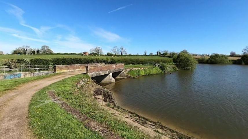 A large reservoir to the right with hedges covering the bank. There is a road with a brick-built bridge to the left.  A field is behind the road to the left and a house is visible on the horizon.