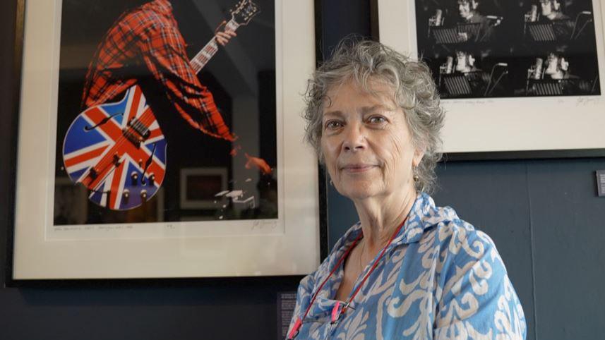A woman with grey hair and blue and white shit. Framed photographs hang on the wall behind her.