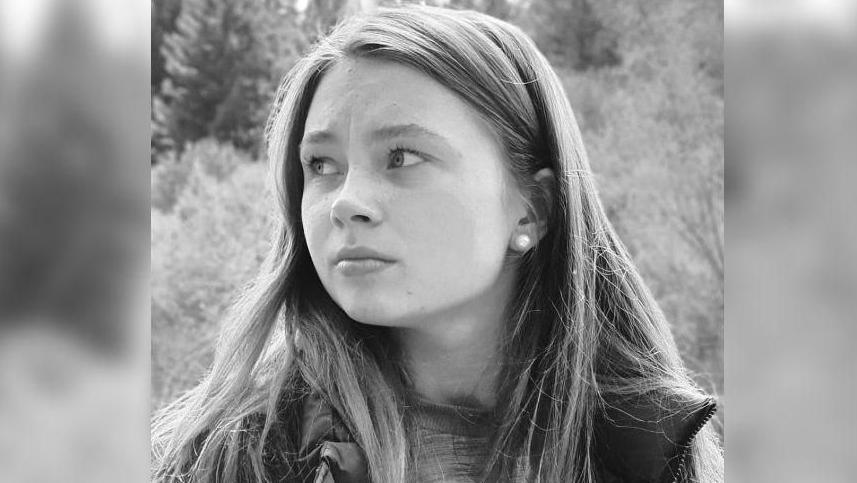 A black and white photo of Jaysley Beck. She is wearing a t-shirt underneath a waterproof jacket, and standing outdoors. Behind her there is an upwards slope of thick trees. She has her long straight her down and is looking off to the left of the camera with a thoughtful expression.