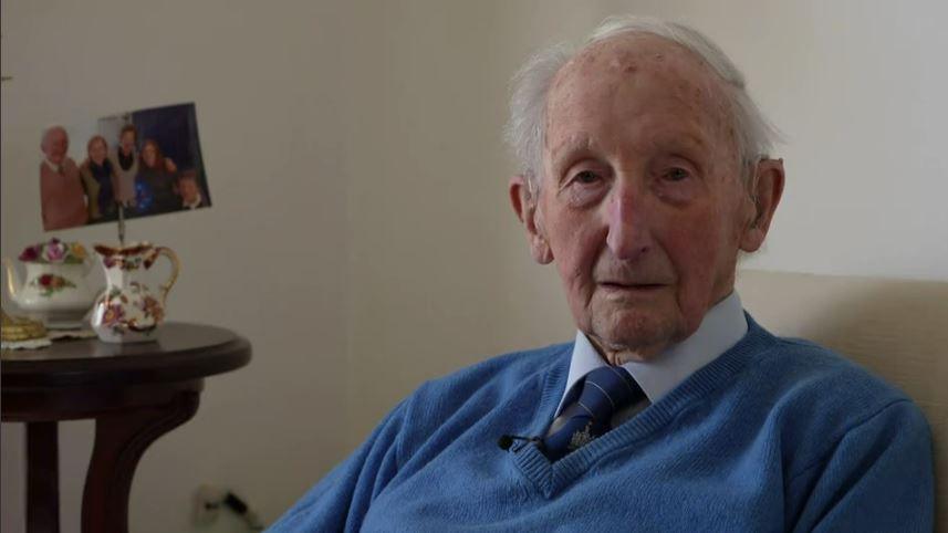 George Young, an older man, looks to the right of the camera, wearing a blue jumper, shirt and tie as he sits in a living room. A table with a family photograph sits beside him to his left.