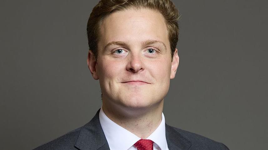 Head and shoulders image of Oliver Ryan standing in front of a dark grey background. He has wavy brown hair and is smiling. He is wearing a dark grey suit, white shirt and red tie