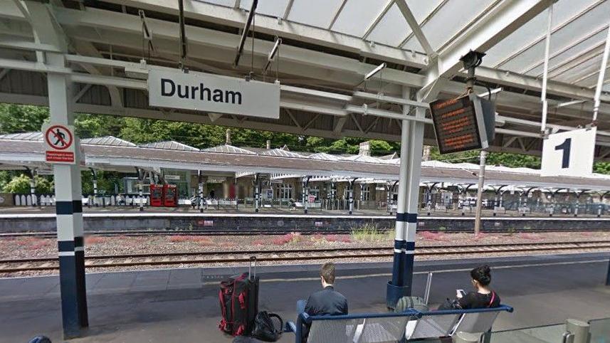 A female and a male passengers sitting on a bench at the southbound Platform 1 at Durham Train station. They are scrolling on their phones, with their suitcases in front of them. There is a sign reading Durham and a screen showing train timings. The Northbound Platform 2 is visible across the tracks.