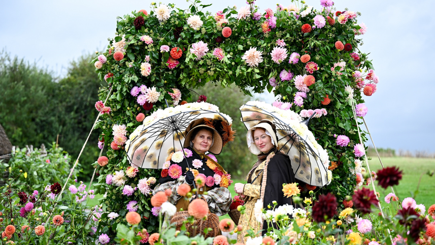 Two ladies dressed in Victorian costume with parasols and surrounded by floral displays and an arch covered in trailing blossoms