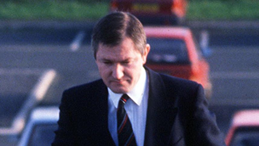 Pat Finucane walking up steps by a car park, he has short brown hair and is staring at the ground. He has a navy suit on with white shirt and black tie with red and grey stripes. 