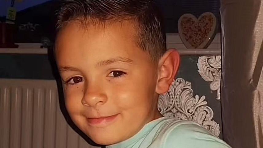 Darrian Williams as a little boy, sitting in a living room in front of a radiator. He has dark hair spiked up in a point and shaved short on the sides. He has small freckles under his eyes and is smiling at the camera, wearing a light blue t-shirt.