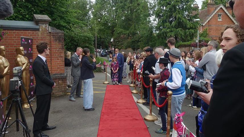A group of people are stood behind a red rope as they are about to watch people walk across the red carpet. One young girl wearing a long purple dress is seen about to walk the red carpet, while on the left there are people wearing suits and smart clothing and a camera.