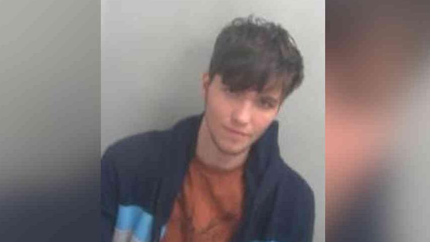 Chase Wiseman has short brown hair with a fringe in his police custody photo. He has a blue jacket and a camel brown T-shirt on.