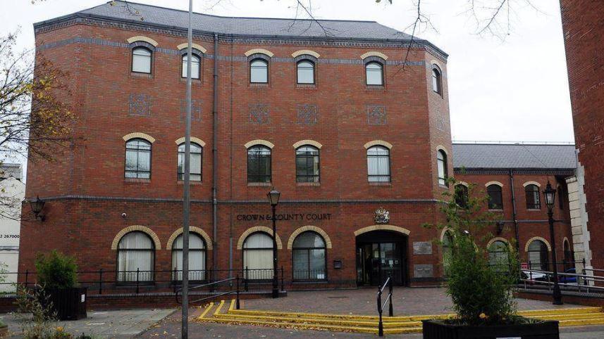 A tall brick building with three rows of art deco-style windows. On the ground are three steps leading to a paved area in front of the entrance.
