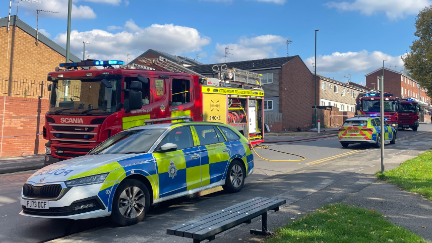 Three fire crews attend blaze at a bungalow in Sneinton, Nottingham. Two police cars are parked on the lerb of a road with fire engines alongside them with brick buildings behind then.