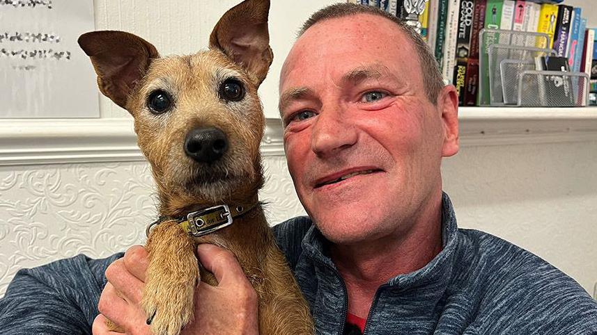 A small, perky, brown and tan jack russell-type dog on the left is looking straight at you. She's being held by Jon in the crook of his arm and shoulder, with his hand supporting her chest. He's in his 50s with blue eyes and short light brown hair. He's clean shaven and is wearing a blue and white marl zip-up hoodie. Behind him is a shelf of books and a notice with the building's wi-fi code, which we've pixelated.