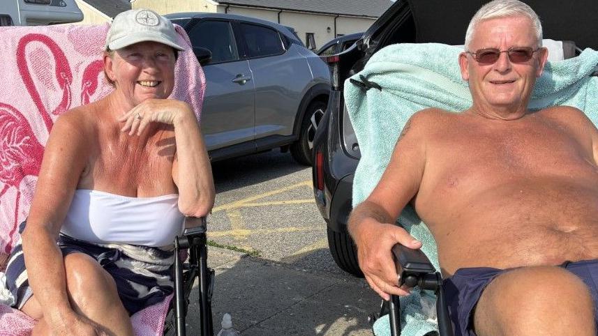 Pauline Stain has a white sunhat and white tokp. Richard HOwared has white hair sunglasseds and blue shnorrts. Both arwe seated in deck chairs.