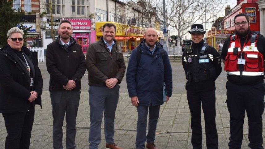 Six people - including a police officer - stand on a paved street with amusement arcades in the background.