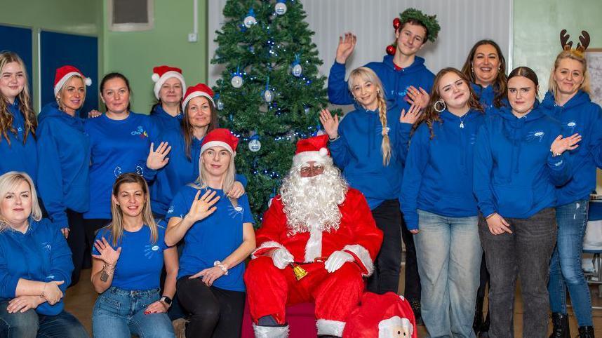 A man dressed as Father Christmas sits in front of a Christmas tree. On either side are young men and women, all wearing a blue top or blue hoody and smiling at the camera. 