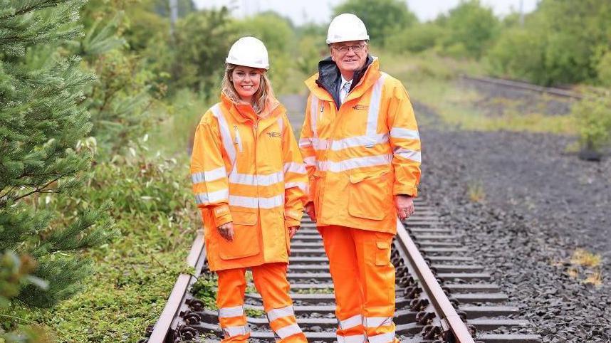 A blonde woman standing next to an older man. Both are wearing orange hi-vis jackets and trousers and hard hats. They're standing on a railway line surrounded by green bushes.