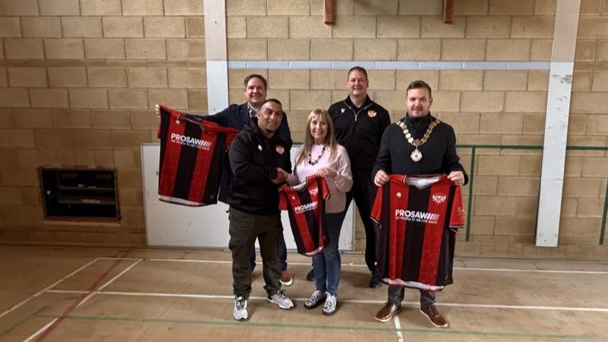 Four men, all dressed in black tops, and a woman, in a pink jumper, stand in a sports hall holding up three red and black football shirts. 