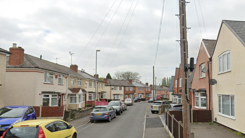 A residential street with cars lining one side. It is a narrow road with houses either side and communication poles can also be seen.