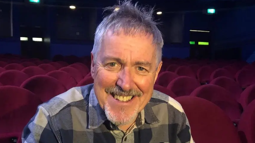 Griff Rhys Jones is sat on a red chair in a theatre. He has short grey hair and is looking towards the camera smiling. He is wearing a grey chequered shirt.