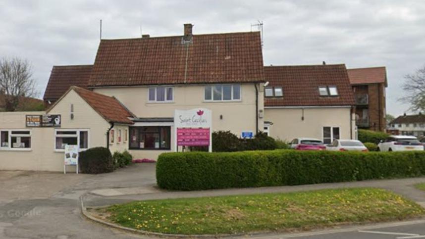 A cream building with red tiled roof surrounded by green privet hedge houses a white and pink wooden sign which reads St Cecilia's Day Centre.