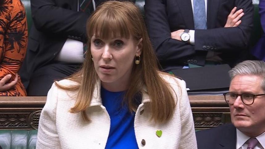 Angela Rayner, a Caucasian woman with red hair wearing a white suit jacket with a green heart-shaped pin and cobalt blue top, speaks in the House of Commons