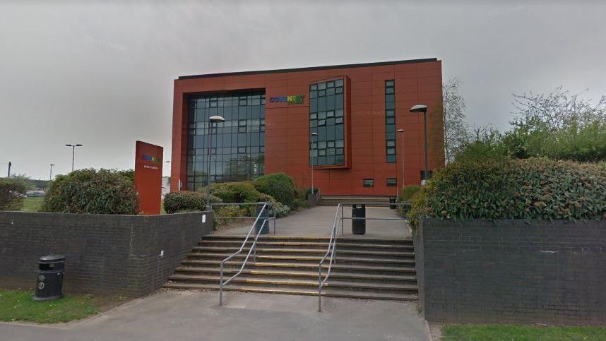 Coventry College's Henley campus, with steps and railings leading to a pathway in front of a college building with red walls and large glass windows. There is a sign on the left hand side and the landscaped area has walls and shrubs.