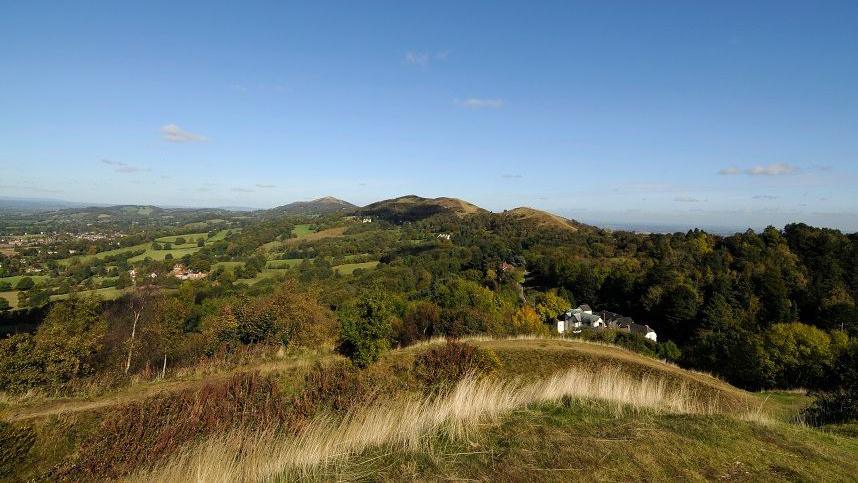 A view from on top of the Malvern Hills on the border of Worcestershire and Herefordshire. There are white buildings in the distance, with hilltops further away, and there is woodland around the houses.