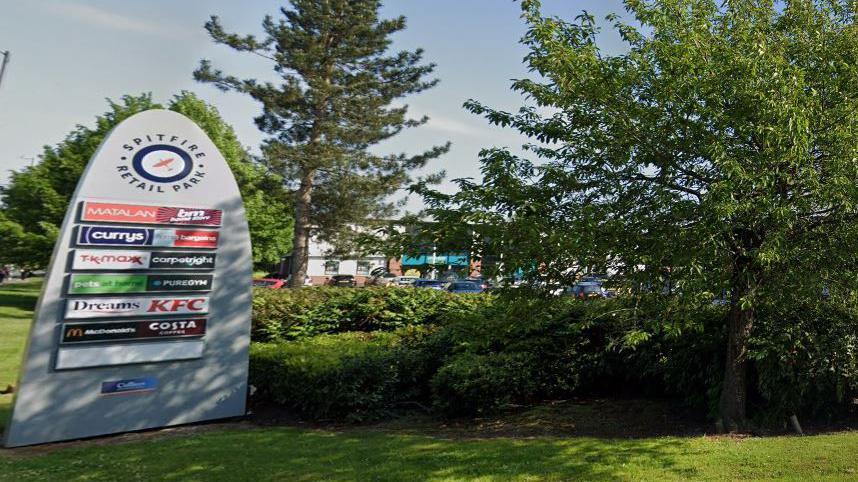 A retail park sign to the left with the logos of the businesses on it sitting in the entrance, surrounded by trees and grass