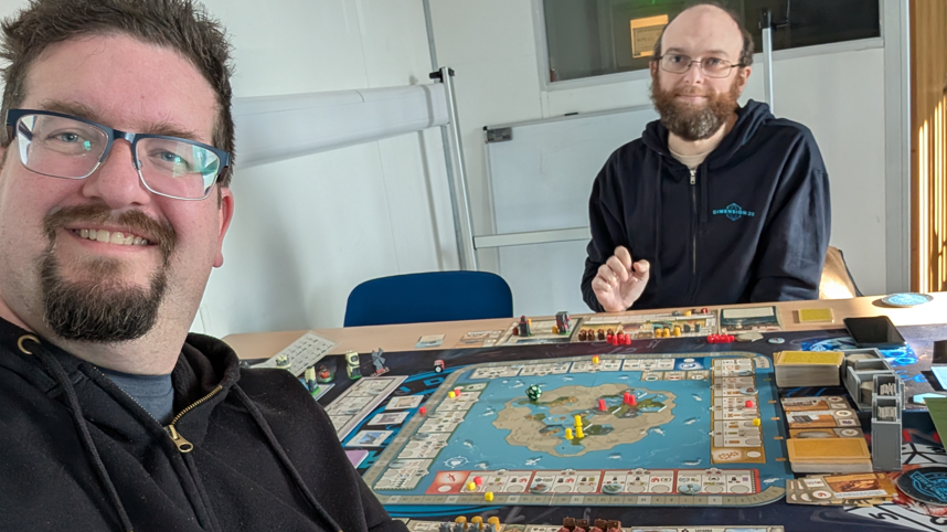 Rikh on the left is taking a selfie with Stuart. They are sitting at a table with a complicated board game at play in front of them