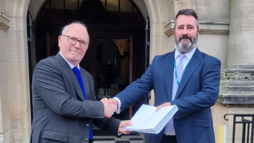 Chris Hall, from SHOUT, hands the petition to Daniel Harry, North Yorkshire Council's head of democratic services. Two men, both wearing suits, light-coloured shirts and tie, shake hands and exchange a paper folder.