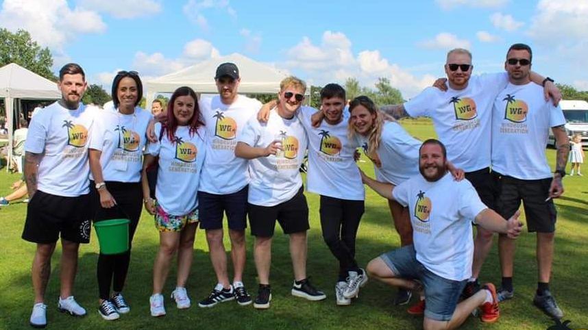 Seven men and three women all wearing the same white t-shirt stand in a park. The T-shirts have a large logo including a sun, palm tree and the letters WG. It is a sunny day and the majority of the group are wearing black shorts and trainers. A gazebo and van can be seen in the background.