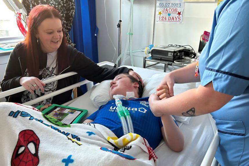 Woman with hand on head of teenage son in hospital bed, with nurse holding his hand