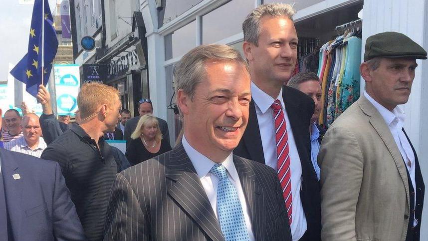 Nigel Farage dhe Nathan Gill ecin në krye të një grupi njerëzish në një rrugë në Caerphilly. Një flamur i BE-së shihet në sfond. Farage është në qendër të fotografisë, Gill është në të majtë të tij dhe pak pas tij. Të dy burrat kanë veshur kostume të errëta, këmisha të bardha dhe kravata.  