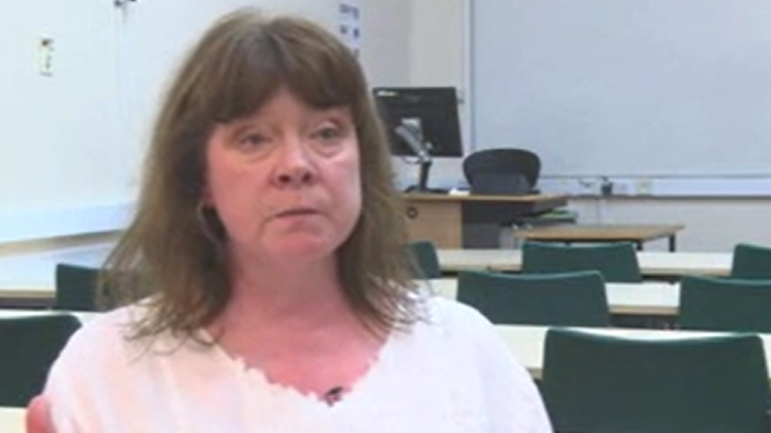 A woman with brown hair, a brown fringe and a white T-shirt sits in a lecture hall in the university. Behind her are desks and chairs pointing towards a whiteboard.