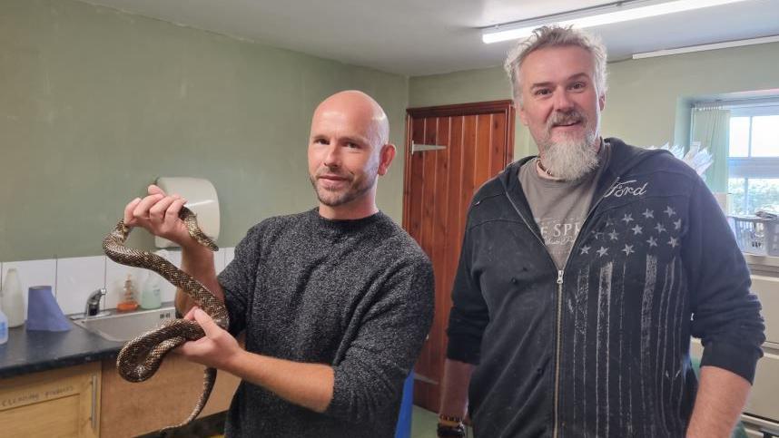 The owner of the snake holding it and looking into the camera with Jamie Mintram standing next to him and smiling in a room with a sink, door and window behind them