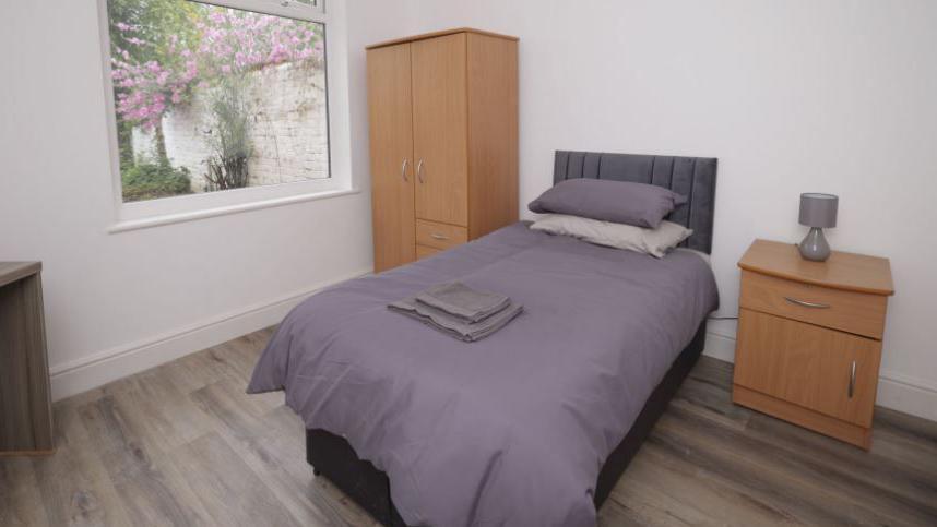 Rehab room with a window overlooking a tree with purple flowers. It has a double bed with a purple duvet with towels folded on the bedspread. It also has a brown wardrobe and bedside table with a purple lamp. 