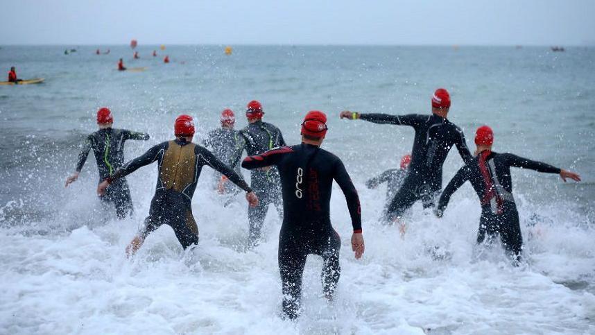 A group of participants wading into the sea wearing wetsuits and red swimming caps as they compete in the swim leg of the race of the IRONMAN 70.3 Weymouth in 2018. People can be seen further out to sea in the distance.