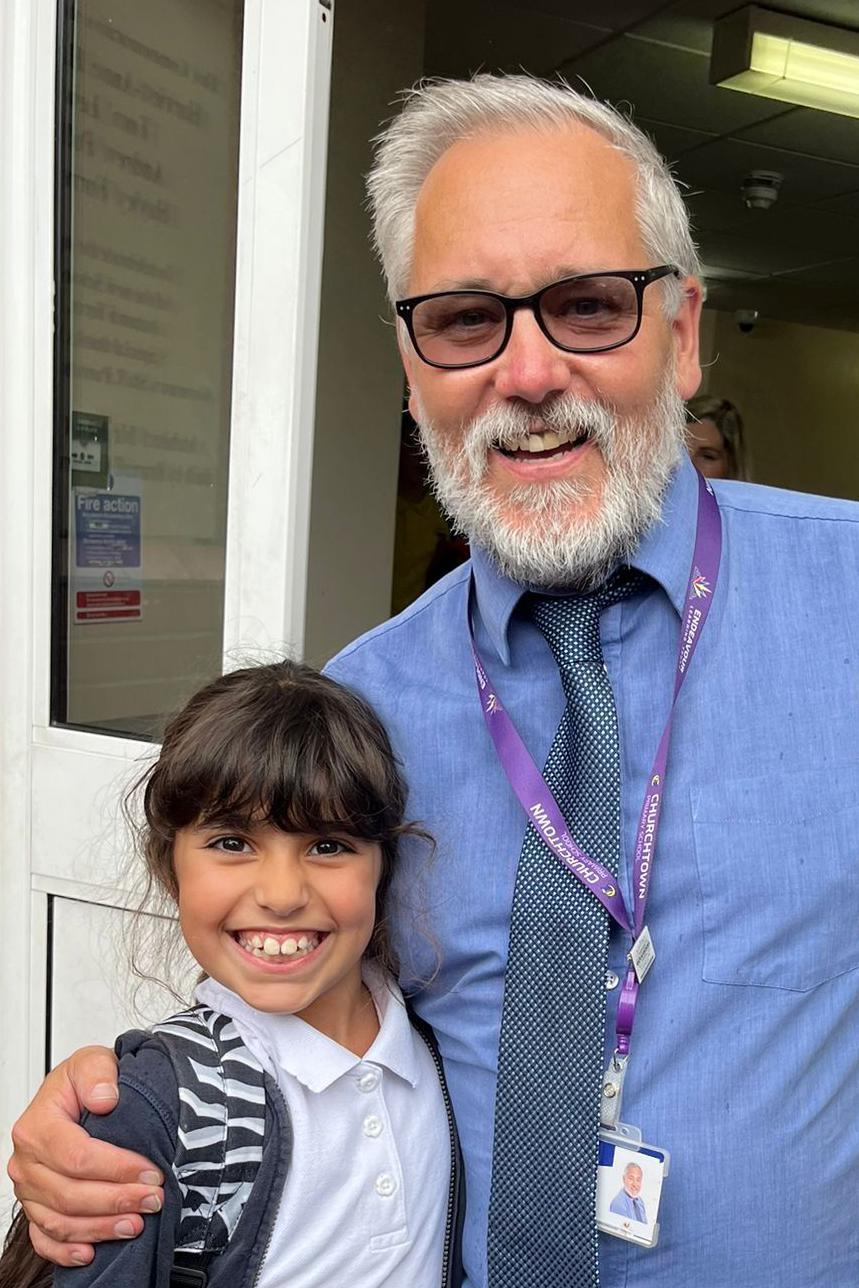 Alice Aguiar smiles at the camera as she stands next to Alan Bowen, her favourite schoolteacher. She is wearing a white polo shirt while he is wearing a blue shirt and spotted purple tie.