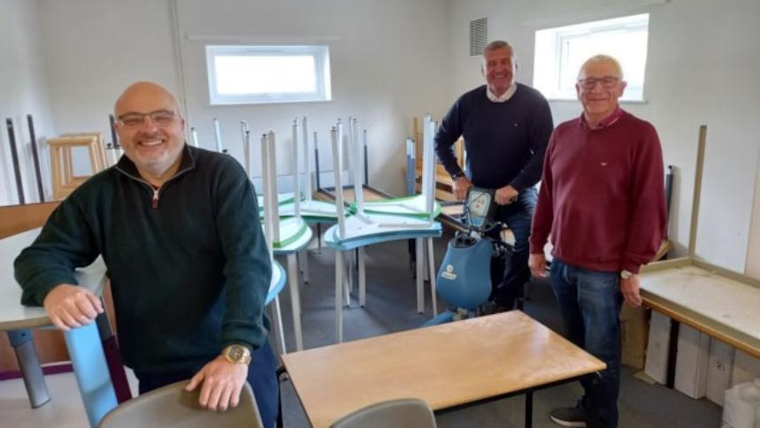 Three male venue trustees smiling while standing in a room at the centre with donated chairs and tables