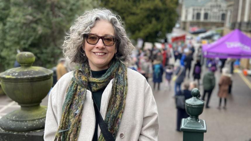 Jane Gill outside the council HQ. She has curly, white hair and glasses. She is wearing a green and yellow scarf and a cream coat. 