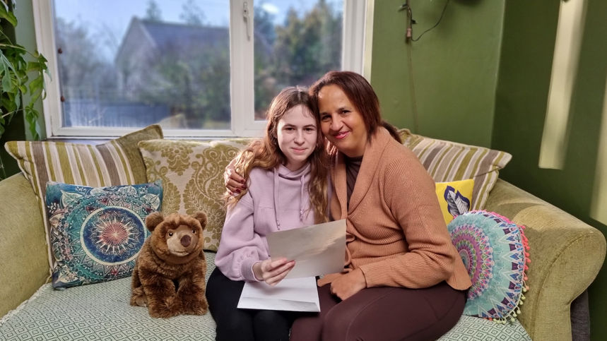 A foster child being hugged by a foster carer on a sofa, smiling to camera and holding a letter