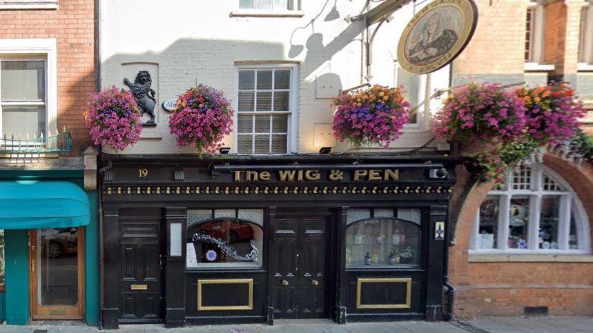 Two-storey pub building with black-painted ground floor entrance, with gold detailing. The words THE WIG & PEN are visible above the door. There are arch-framed windows either side of the door, and purple-flowered hanging baskets above. The first floor is painted white.