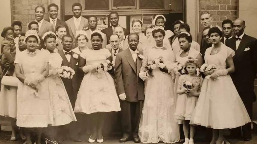 A black and white image of a bride and groom surrounded by other men and women in white dresses and tuxedos. The women are holding floral bouquets.