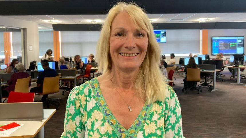 A woman in a green floral dress with long blond hair, standing smiling in a call centre.