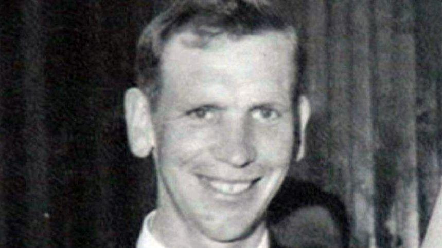 The image is black and white and shows the head and shoulders of a man. He has short hair and is smiling towards the camera. 