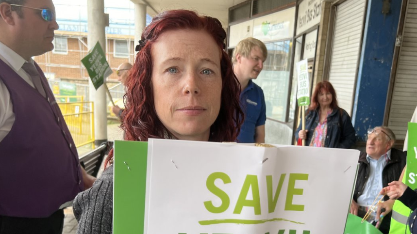 Linda Snelling from Yeovil's bus users group.  The photo shows Linda at a previous demonstration to save Yeovil bus station. Linda is holding up a green and white protest banner and is standing in front of a group of people.