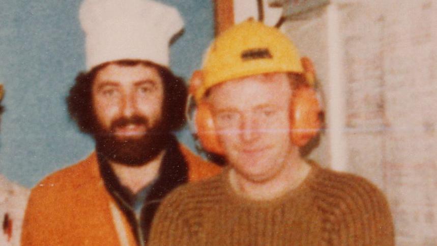 Old picture of two men standing side by side looking at the camera. The man on the left is wearing an orange boiler suit, a white chef's hat and has a thick black beard. The one on the right has an orange jumper, a yellow helmet and orange wear defenders.