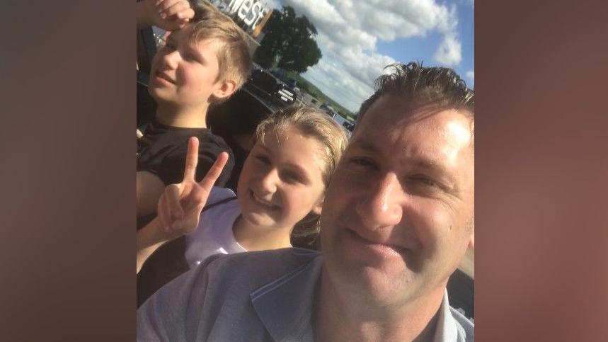 Nik Hirst, pictured in a selfie at a fair with his youngest children, one child is making the peace sign with their fingers.