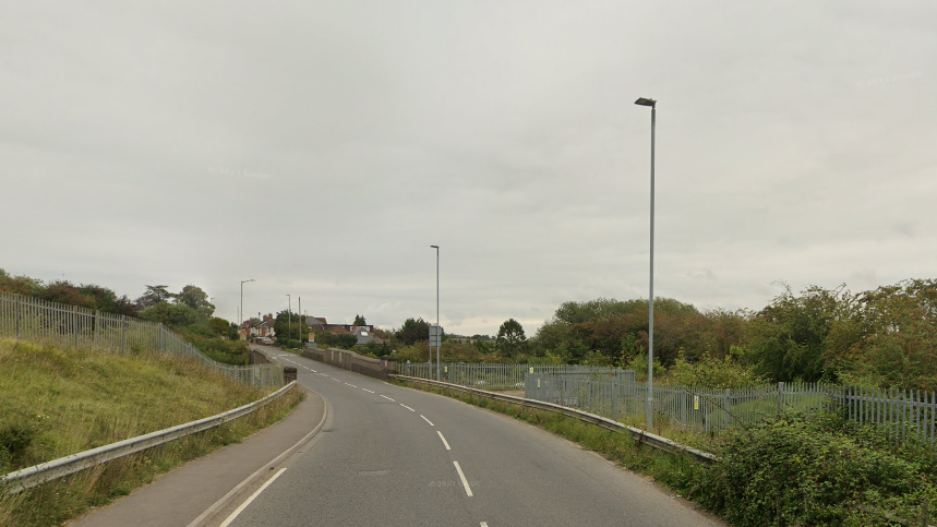 View of A3102 leading into Royal Wootton Basset. There are bushes on the right and the sky is grey.